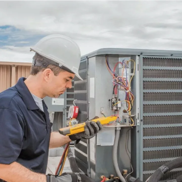 HVAC technician in a hard hat testing an outdoor air conditioner with a multimeter